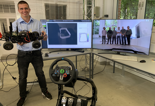 Group picture. One member standing in the driving central, the others are viewed by the car on the racetrack and are displayed on screen